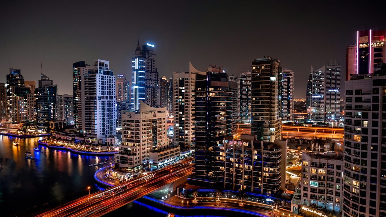 Night view of Dubai Marina’s high-rise skyline, with traffic light trails on a bridge and reflections in the canal