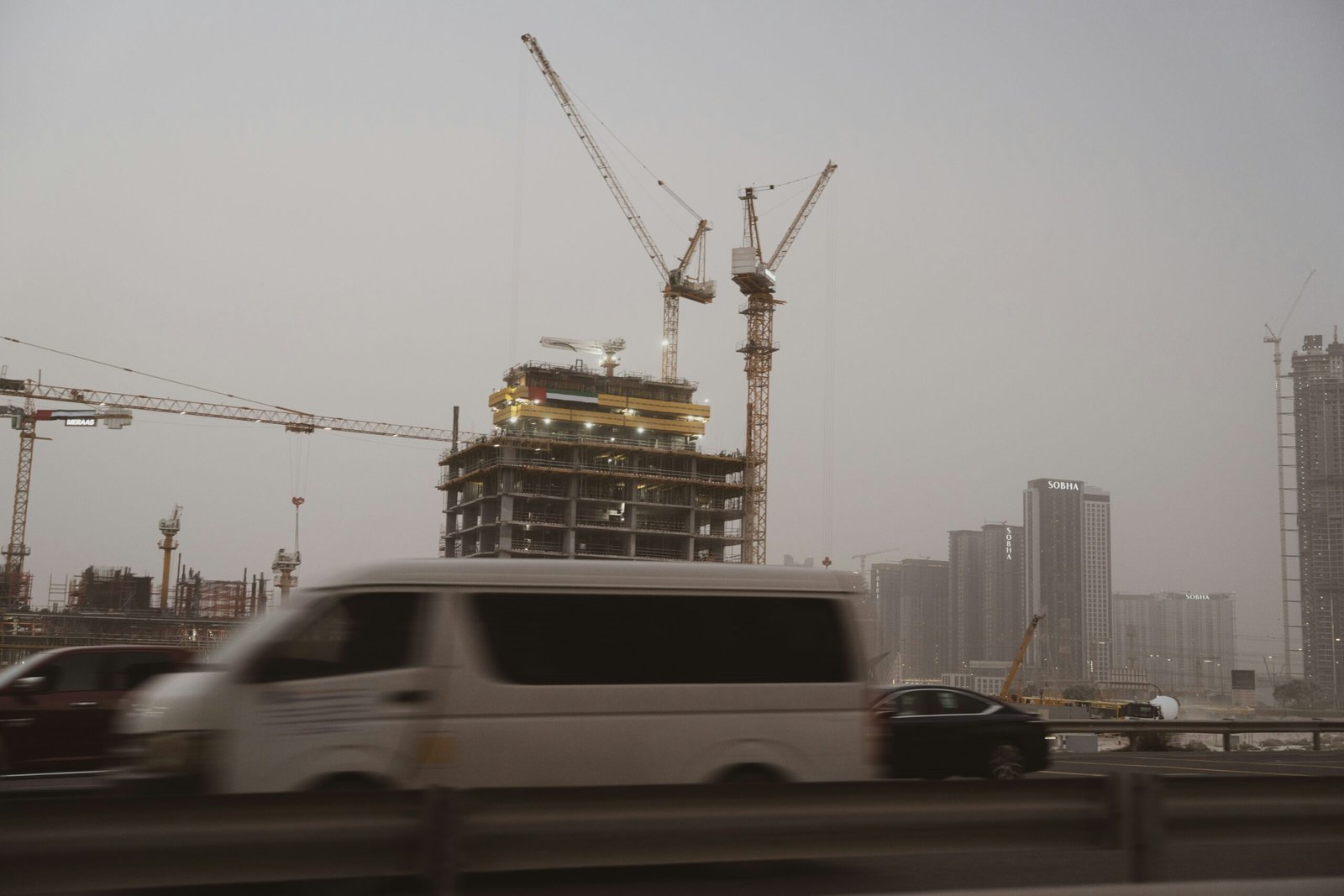 Construction cranes and a high-rise building under development beside a busy road in Dubai, with vehicles passing in the foreground.