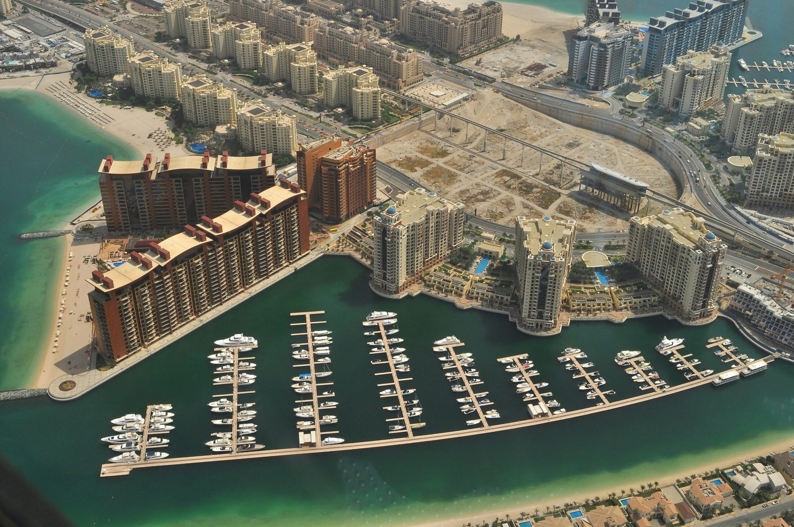 Aerial view of a Dubai waterfront marina with residential towers, private docks, yachts, and sandy shoreline.