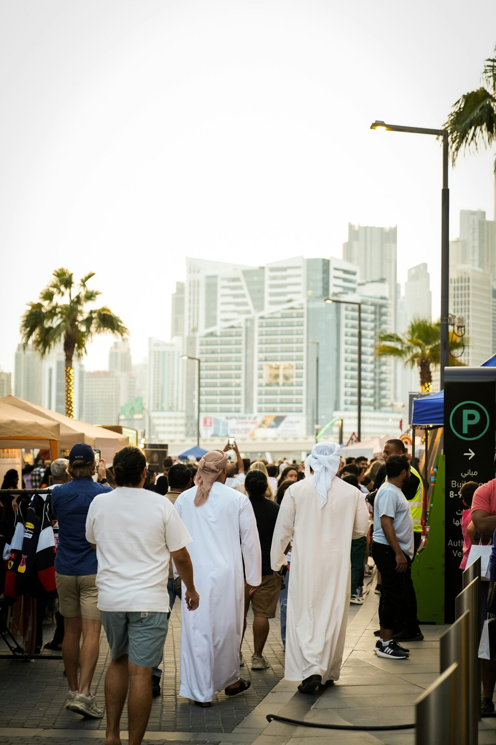 People walking through a busy public area in Dubai, with a diverse crowd, palm trees, and high-rise buildings in the background.
