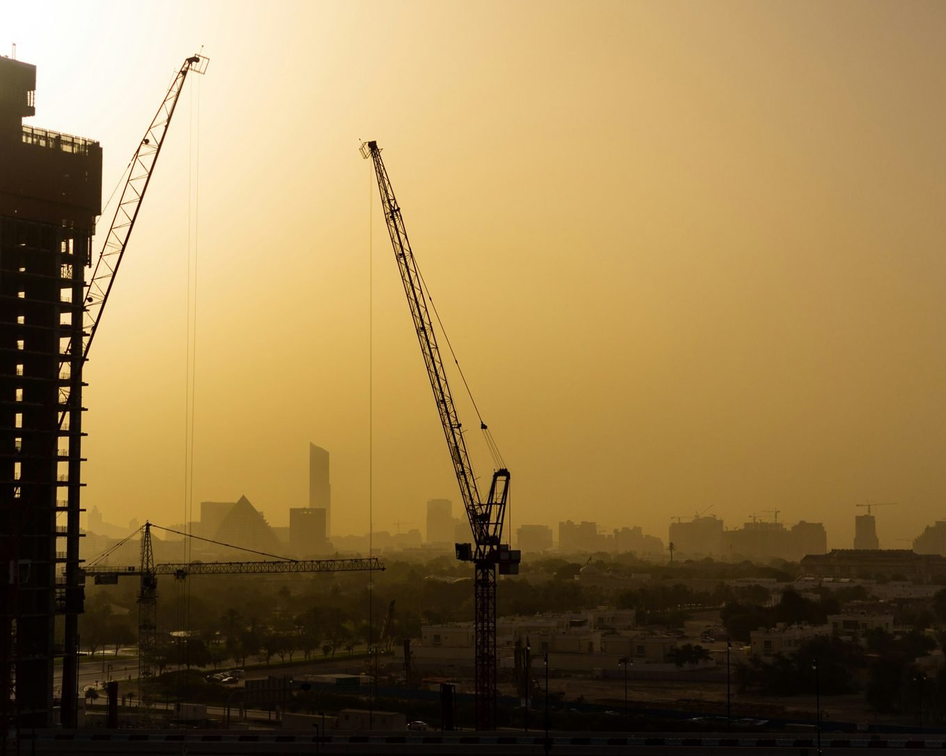 Two tall construction cranes beside a high-rise under construction, with a hazy city skyline in the background at golden hour, reflecting the off-Plan Property in Dubai article.
